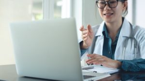 Asian,Doctor,Using,Laptop,Computer,Online,Video,Call,,Telemedicine,To Sick girl wrapped in scarf holding tissue at face and consulting doctor online on video call
